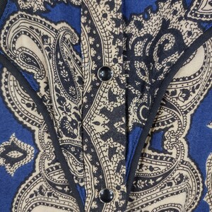 May include: A blue and beige paisley patterned shirt with a black trim and two black buttons.