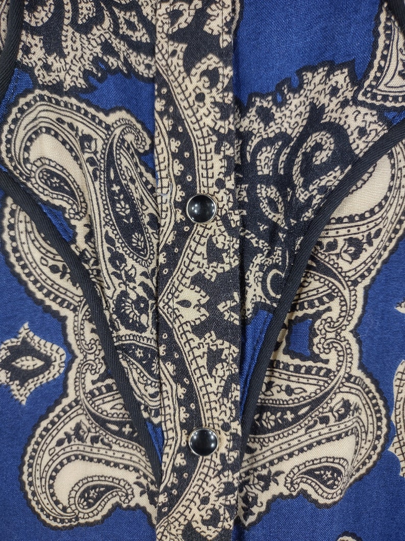 May include: A blue and beige paisley patterned shirt with a black trim and two black buttons.