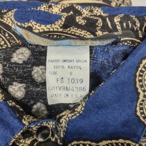 May include: A blue and beige paisley patterned shirt with a white tag that reads "Fabric: Import Origin 100% Rayon Size S FS 1039 LOTV8M4386 Made in U.S.A."