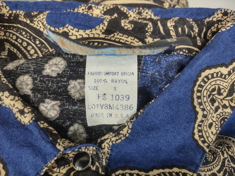 May include: A blue and beige paisley patterned shirt with a white tag that reads "Fabric: Import Origin 100% Rayon Size S FS 1039 LOTV8M4386 Made in U.S.A."