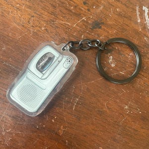 Könnte beinhalten: Ein durchsichtiger Schlüsselanhänger aus Kunststoff in Form eines Vintage-Kassettenrekorders. Der Schlüsselanhänger hat eine schwarze Kette und einen silbernen Ring. Der Kassettenrekorder trägt die Aufschrift "PLAYTIME".