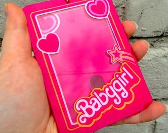 May include: A rectangular hot pink acrylic card holder featuring a clear central window. The frame is decorated with pink and white heart graphics, a shooting star, and the word "babygirl" in white script with a yellow and orange outline.