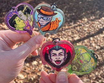 May include: Four heart-shaped keychains with different monster characters. The keychains are made of clear plastic with a colorful design. The characters include Frankenstein's monster, the Creature from the Black Lagoon, Dracula, and the Invisible Man.