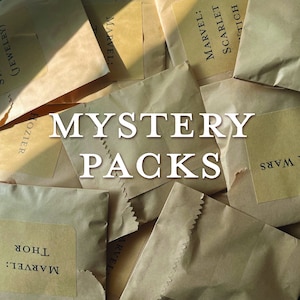 May include: A close-up of a pile of brown paper bags with different text on them. The text includes "MARVEL", "SCARLET", "WARS", "THOR", and "HOZIER". The text "MYSTERY PACKS" is written in large white letters across the center of the image.