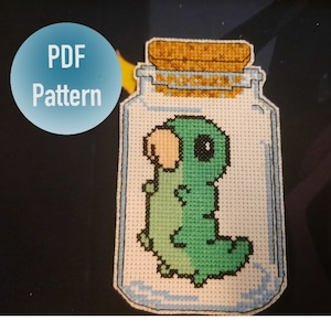 Pode incluir: Um padrão de ponto cruz de um verme verde em um frasco de vidro com tampa de cortiça. O verme tem um corpo verde claro e uma cabeça verde escura. O frasco é transparente e tem uma borda azul.