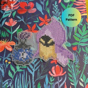 May include: A purple and yellow cross-stitch pattern of a hooded figure with a black eye. The pattern is on a white background and has the text "PDF Pattern" in a blue circle.