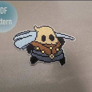 May include: Cross-stitch pattern of a beige and brown character with a blue bag, a brown hat, and a black and blue outfit. The character is holding a sword in its right hand. The pattern is labeled "PDF Pattern".