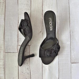Op de afbeelding: Een paar zwarte satijnen sandalen met hak en een bloemversiering op de teen. De sandalen hebben een lage hak en zijn op de binnenzool gemarkeerd met "Moda".