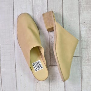 May include: A pair of light beige leather mules with a block heel, set against a light wood-grain background. One mule is a closed-toe design, while the other is open-backed. The shoes have a minimalist aesthetic, suitable for casual wear.
