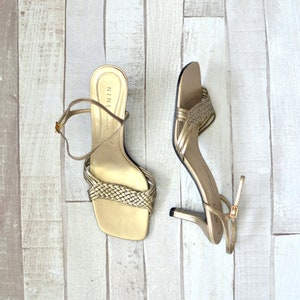 May include: A pair of gold-colored Nine West heeled sandals. One sandal has a woven strap across the toes, and the other has multiple thin straps. Both have a thin ankle strap with a buckle. The heels are approximately 3 inches high.