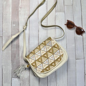 Op de afbeelding: Crèmekleurige crossbodytas met een geometrisch patroon van gouden, zilveren en beige driehoeken. De tas heeft een lange, dunne riem en een kwast. Bruine zonnebril ligt ernaast op een houten oppervlak.