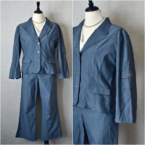 Könnte beinhalten: Ein denimblauer zweiteiliger Anzug auf einer Schaufensterpuppe. Der Anzug besteht aus einem Blazer mit zwei Vordertaschen und einer Hose mit weitem Bein. Der Blazer hat Knopfverschlüsse und hochkrempelbare Ärmel. Ein weißes Oberteil und eine goldene Halskette sind sichtbar.