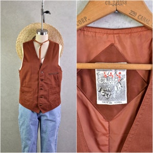 May include: A brown vest with a V-neck, button closure, and two front pockets. The vest has a small pocket on the chest and is displayed on a mannequin. The vest is paired with blue jeans and a straw hat.