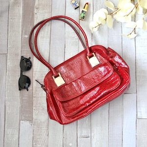 May include: A red handbag with a crocodile skin texture, gold-colored hardware, and a front pocket. Black sunglasses and two tubes of lipstick are next to the bag. The bag has two handles.