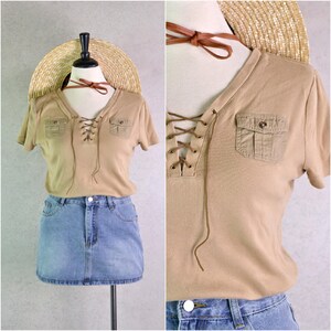 May include: Tan short-sleeve shirt with a V-neck and lace-up detail. Features two small pockets and is paired with a denim skirt and a straw hat. A brown leather choker is also included.