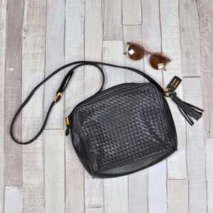 Könnte beinhalten: Schwarze Leder-Umhängetasche mit gewebter Front, langem Riemen und Quaste. Die Tasche hat goldfarbene Hardware und ein Etikett mit der Aufschrift "Genuine Leather". Eine braune Sonnenbrille liegt daneben.