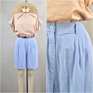 Puede incluir: Un atuendo vintage con una blusa color melocotón con cuello en V y mangas enrolladas, combinada con pantalones cortos azul claro de cintura alta. Un cinturón marrón y un sombrero de paja completan el look. Los pantalones cortos tienen un diseño plisado.