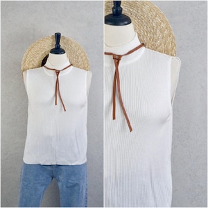 Puede incluir: Top sin mangas blanco acanalado con detalle de lazo de cuero marrón. El top se muestra en un maniquí con un sombrero de paja en el fondo. El top se combina con unos vaqueros azules.