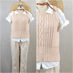 Puede incluir: Un chaleco de punto de cable beige claro con un diseño de cable central y un dobladillo acanalado. Se superpone a una camisa blanca con cuello con mangas enrolladas y se combina con pantalones beige. También se ve un sombrero de paja y un cordón de cuero marrón.