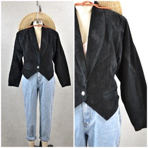 May include: Black suede blazer with a single button closure and a notched lapel. The blazer is paired with light wash denim jeans. A straw hat is visible in the background.