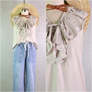 May include: A beige sleeveless top with ruffled detailing and a V-neck, paired with light blue denim pants. A straw hat and a brown leather neck tie are also visible. The outfit is displayed on a mannequin.