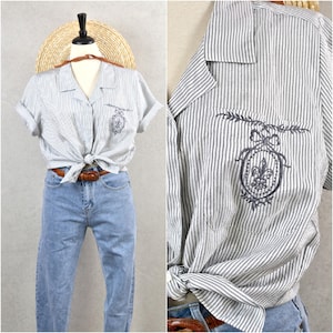 Puede incluir: Camisa a rayas blancas y grises de manga corta, anudada a la cintura, combinada con vaqueros azul claro. La camisa tiene un emblema bordado en el bolsillo. Un cinturón marrón y un sombrero de paja completan el conjunto.