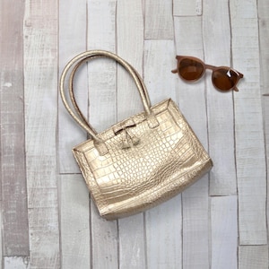 May include: A gold-colored tote bag with a crocodile skin texture, featuring two handles and a decorative tassel. Brown sunglasses are placed next to the bag. The bag is made of a shiny material.