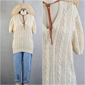 Puede incluir: Suéter de punto trenzado color crema con mangas cortas y cuello en V profundo. El suéter se combina con jeans azul claro. Se presenta una corbata de cuero marrón. Un sombrero de paja se coloca encima de la cabeza del maniquí.