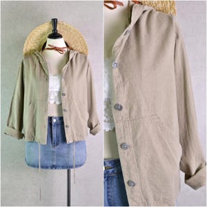 May include: A tan hooded jacket with button closures and front pockets. The jacket is paired with a denim skirt and a straw hat. The jacket's sleeves are rolled up, and the fabric appears to be linen or a similar lightweight material.