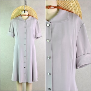 Puede incluir: Un vestido lila claro con mangas cortas y botones en la parte delantera. El vestido tiene un cuello y una falda acampanada. Los botones son nacarados. Un sombrero de paja está colocado en el maniquí.