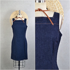 Puede incluir: Vestido sin mangas de mezclilla azul oscuro, con escote cuadrado y silueta entallada. El vestido se exhibe en un maniquí con un sombrero de paja y una correa de cuero marrón. El vestido tiene costuras visibles y una abertura trasera.
