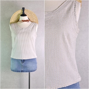 Puede incluir: Un top sin mangas beige y blanco texturizado con cuello redondo y bordes deshilachados. El top se combina con una minifalda vaquera. También se muestra un sombrero de paja y un collar de cuero marrón.