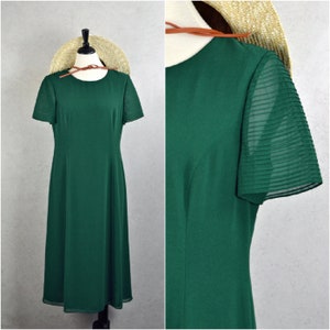 Puede incluir: Vestido verde oscuro con mangas cortas plisadas y cuello redondo. El vestido se exhibe en un maniquí con un sombrero de paja y una correa de cuero marrón. El vestido tiene una falda fluida de largo midi.