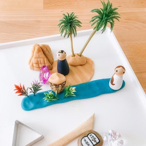 May include: A play scene with a blue river, two palm trees, a brown figure, a brown pyramid, a basket, and a pink gem.