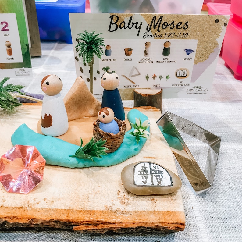 May include: A wooden playset depicting the story of baby Moses in a basket, with a brown, tan, and blue wooden figures, a green plant, a blue play dough river, and a stone with hieroglyphics. The playset is on a wooden base.