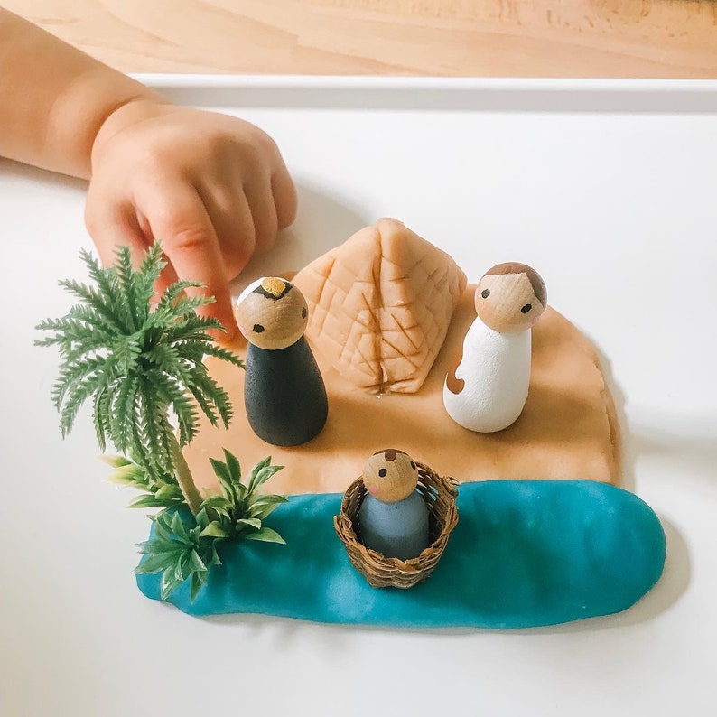 May include: A playset featuring a brown pyramid, a blue river, and three wooden figurines. The figurines are dressed in white, black, and blue. The figurines are placed on a brown play dough surface.
