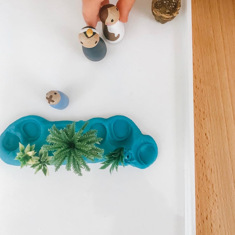 May include: A blue play dough mat with five indentations, resembling a pond, with three small green plants and a wooden figurine of a person.