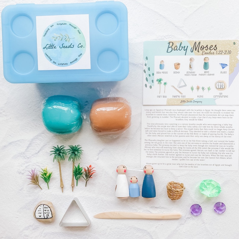 May include: A blue plastic storage container with a label that reads "Little Seeds Co. Teaching the scripture through play." The container is open and contains a set of play dough, wooden figurines, a small wooden basket, a wooden stick, a small wooden boat, a small wooden palm tree, a small wooden stone, a small wooden triangle, and a small wooden stone with the text "VII" on it. The set is designed to help children learn about the story of Baby Moses from the Bible.