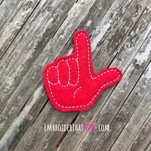 May include: Red felt hand with white stitching, making the "I love you" hand gesture. The hand is on a wooden background.