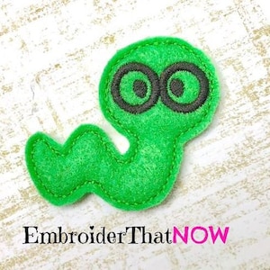 Könnte beinhalten: Ein hellgrünes Filz-Applikation in Wurmform mit großen, schwarz umrandeten Augen. Die Applikation ist mit passendem grünen Faden genäht. Der Text "EmbroiderThatNOW" ist darunter in Schwarz und Pink gedruckt.