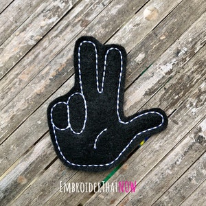 以下が含まれることがあります： 白いステッチで縫われた黒いフェルトの手のアップリケで、ピースサインのジェスチャーで3本の指を立てています。