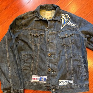 May include: A dark wash denim jacket with a button-up front, two chest pockets, and two side pockets. The jacket features a team logo patch on the upper left chest and CCM and NHL patches. The jacket is a classic style.