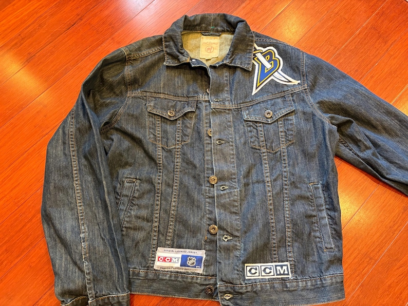 May include: A dark wash denim jacket with a button-up front, two chest pockets, and two side pockets. The jacket features a team logo patch on the upper left chest and CCM and NHL patches. The jacket is a classic style.