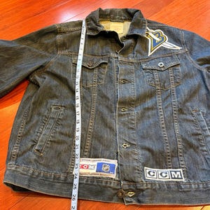 May include: A dark blue denim jacket with a button-up front, pockets, and a collar. Patches include a logo with a yellow and blue design and the letters "CCM". The jacket is approximately 26 inches long.