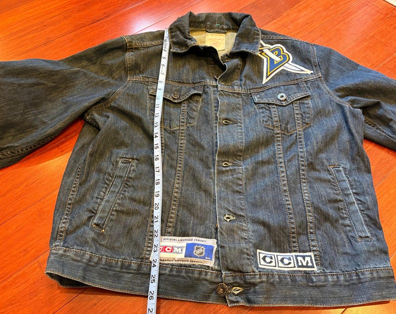 May include: A dark blue denim jacket with a button-up front, pockets, and a collar. Patches include a logo with a yellow and blue design and the letters "CCM". The jacket is approximately 26 inches long.
