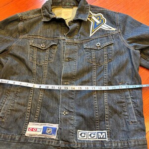 May include: A dark wash denim jacket with a button-up front, chest pockets, and a collar. The jacket features a patch with a logo and the text "CCM". The jacket measures approximately 24 inches across the chest.