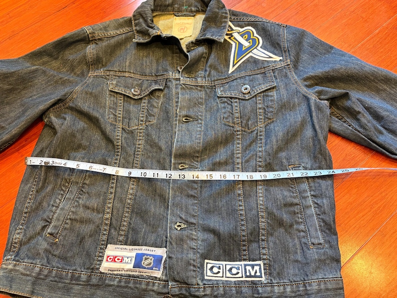 May include: A dark wash denim jacket with a button-up front, chest pockets, and a collar. The jacket features a patch with a logo and the text "CCM". The jacket measures approximately 24 inches across the chest.
