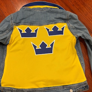 May include: A blue denim jacket with a yellow fabric panel on the back. The panel features three white outlined blue crowns.