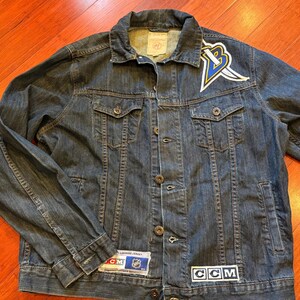 May include: A dark wash denim jacket with a button-up front, two chest pockets, and two side pockets. The jacket features a logo patch on the chest and a CCM patch. A team logo is on the upper left chest.