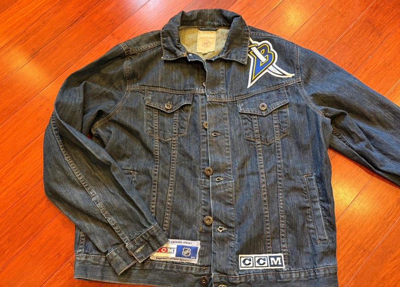 May include: A dark wash denim jacket with a button-up front, two chest pockets, and two side pockets. The jacket features a logo patch on the chest and a CCM patch. A team logo is on the upper left chest.